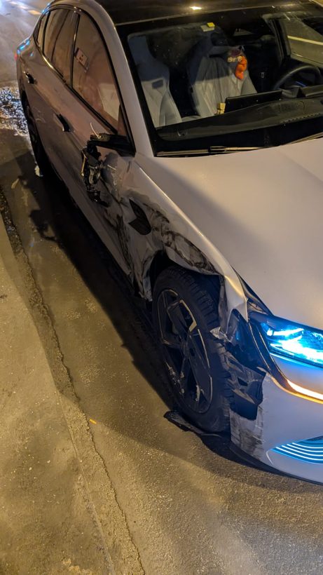 Foto reale dell'auto elettrica del fondatore di Nagi dopo l'incidente notturno in superstrada: fiancata e ruota anteriore sinistra danneggiate dall'urto contro un camion, la firma luminosa teal del faro a LED ancora accesa nella notte.